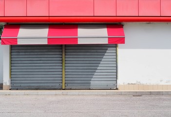 Shop retail with metal shutters closed and a red white awning. Sidewalk and asphalt road in front....