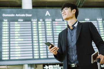 Fototapeta premium Elegant asian businessman checking email on mobile phone while walking with suitcase inside airport, experienced male employer using cell telephone while waiting for taxi car coming before work travel