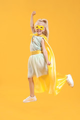 Jumping little girl dressed as superhero on color background
