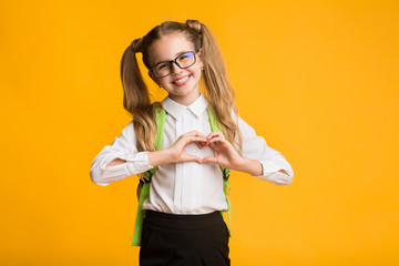 Little smiling schoolgirl showing heart with hands on yellow background