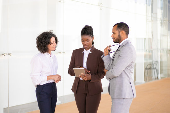 Young female sales agent presenting project to customers. Multiethnic business man and women standing in hallway, using tablet, discussing issues. Partnership concept