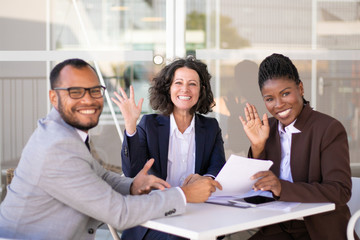 Happy successful business team posing at table and waving hello. Cheerful multiethnic man and women...