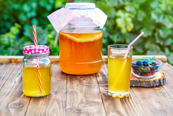Glass jar with kombucha, a poured glass with kombucha and a cup with blackberry in a summer garden.