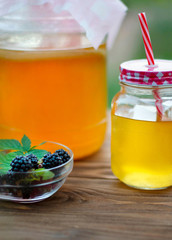 Glass jar with kombucha, a poured glass with kombucha and a cup with blackberry in a summer garden.