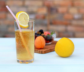 Poured glass with kombucha with a slice of lemon, fruits on a wooden board on a table on a colored brick background in a summer garden.