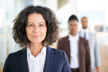 Successful female leader posing with her team in blurred background. Middle aged businesswoman smiling at camera, her two colleagues standing behind her. Successful team leader concept - Powered by Adobe