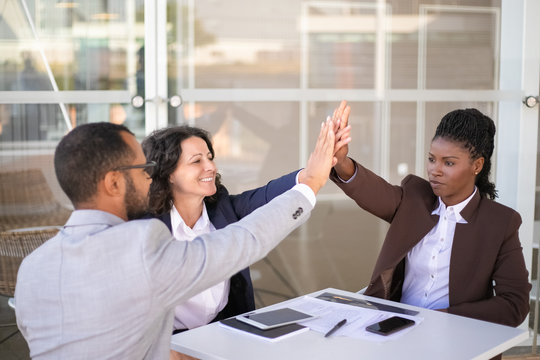Happy businesspeople celebrating team success. Multiethnic group of man and women sitting at table near office building and giving high five to each other. Teamwork concept