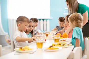 Kids have a dinner in kindergarten. Little boys and girls from the group of children sitting at table with lunch and eat appetizing. Children with caregiver in day care centre. Childs have healthy - 285201587
