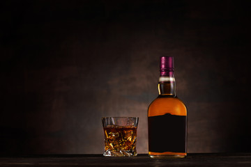 Glass of whiskey with ice and bottle on dark wood background