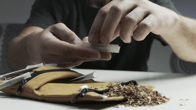 Pan shot of man's hands detail while rolling up a cigarette, tobacco holder and tabacco accessories on the table foreground 