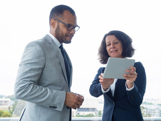 Satisfied business leaders watching project presentation on tablet. Business man and woman standing indoors at office window, woman holding tablet and talking. Business communication concept