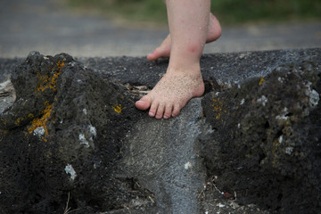 Toddler climbing down rock wall at beach with sandy feet. Could be boy or girl, gender neutral. Rocks have moss and lichen, rustic. Pt Chevalier beach, Auckland. Innocent fun, kiwi kid, nostalgic.