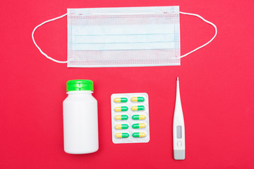 Red desk, medical mask, pills in blister pack, bottle with pills, thermometer, top view, red background