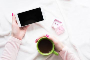 Women's hands holding cup of coffee and smart phone on white plaid, pink box, copy space, top view