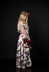Obraz na płótnie Canvas full length portrait of a blonde girl wearing a long white and floral dress, wearing a flower crown. Standing pose on a black studio background.