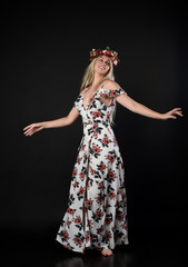 full length portrait of a blonde girl wearing a long white and floral dress, wearing a flower crown. Standing pose  on a black studio background.