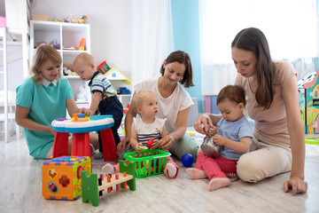 Adorable toddlers playing with colorful toys and mothers in nursery room. Nursery babies playing with adults in daycare center