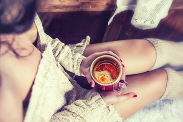 A girl in a warm knitted jacket with long legs sits and holds a red mug with cooling coffee. The concept of Christmas, New year, warm cozy home and melancholy. The view from the top
