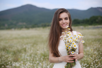 Beautiful girl outdoors with a bouquet of flowers in a field of white daisies,enjoying nature. Beautiful Model with long hair in white dress having fun on summer Field with blooming flowers,Sun Light.