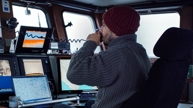 Captain of Commercial Fishing Ship Surrounded by Monitors and Screens Using Radio for Communication and Working with Sea Maps in his Cabin.