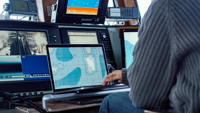 Captain Of Commercial Fishing Ship Surrounded By Monitors And Screens Working With Sea Maps In His Cabin.