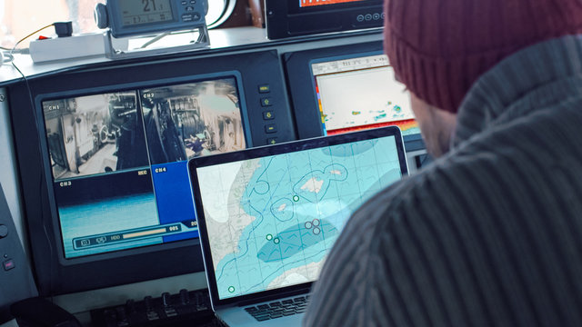 Captain Of Commercial Fishing Ship Surrounded By Monitors And Screens Working With Sea Maps In His Cabin.