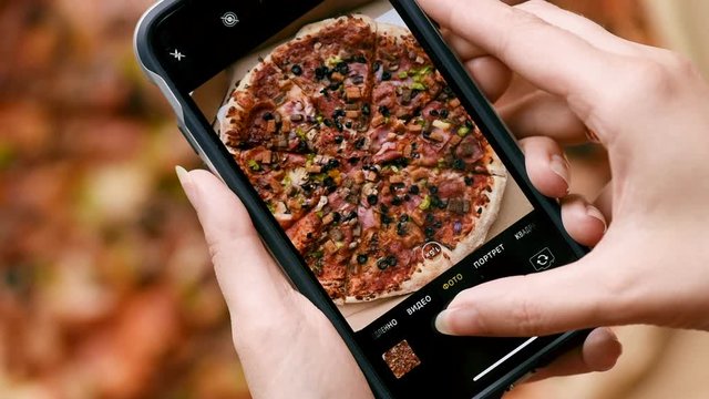 Female hands taking photo of delicious Italian pizza on smartphone. Woman in restaurant make photo of food on mobile phone camera. Top view close-up. People and food concept.