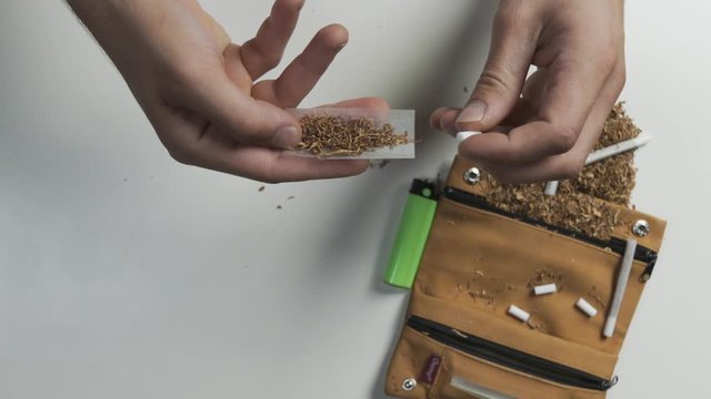 Top view/Zenital shot of man's hands detail showing how rolling up a cigarette, tabacco holder and tabacco accessories on the table background 
