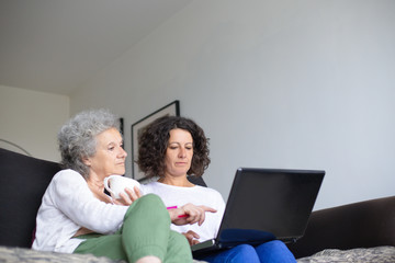 Senior mother with adult daughter using laptop. Low angle view of elderly woman sitting on couch with middle aged daughter and using laptop computer together. Technology concept