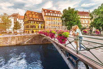 Two young happy girl friends standing on a bridge in Strasbourg while travelling in Petit France region. Tourism and friendship concept - obrazy, fototapety, plakaty
