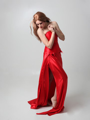 Fototapeta premium full length portrait of a girl wearing a long red silk gown, Standing pose on a grey studio background.