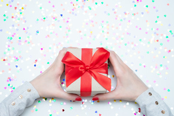 White desk, sparkles hearts, girl holds gift, woman's hands, gift box with red ribbon, close up, copy space