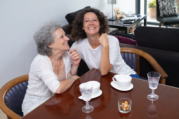 Happy mother and daughter laughing at home. Cheerful senior mother and middle aged daughter sitting at table with cups and glasses. Spending time together concept