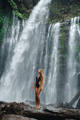 Woman enjoying near hidden in jungle huge waterfall in Indonesia. Slim body and black swimsuit, fashion model. Travelling  Indonesia