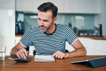 Smiling man managing home budget with calculator and notebook while sitting at dining table