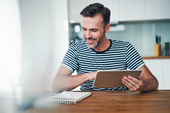 Smiling man sitting at home dining table and using tablet managing personal finances