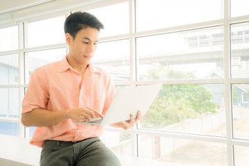 A Asian Handsome businessman 24s working with laptop near window in workplace., A handsome 24 year old Asian male student is learning use a laptop on the table in the home. back to school
