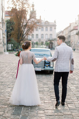 Pretty bride and handsome groom walking in the old city center, standing in front of the black retro car