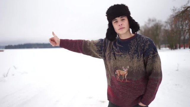 A handsome man in a hat with earflaps hitchhiking on the road. A man stands alone on the road in winter. A man in a hat with earflaps hitch a ride into the woods.
