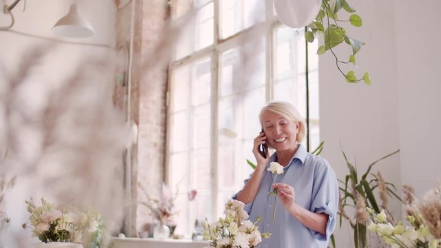 Tilt down of middle-aged blonde woman standing at wooden table, smelling flowers, arranging bunch and having phone discussionTilt down of middle-aged blonde woman standing at wooden table, smelling fl