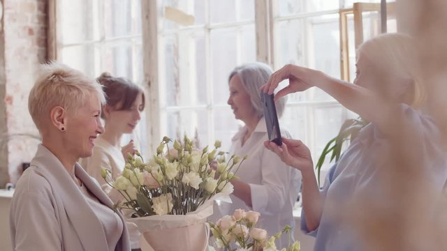 Medium shot of two middle-aged women on foreground making photos with flowers on smartphone while two other women on background talking