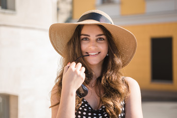 Pretty girl in a black dress and a hat with wide flaps smiles holding sun glasses in teeth enjoying walk down the European town during sunny summer day.