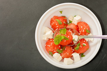 Salad of watermelon balls, feta cheese and mint, sprinkled with sesame seeds in a white plate with a spoon on a black background. Flat lay