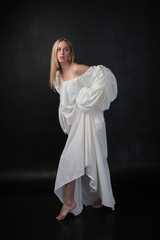 full length portrait of blonde girl wearing long white flowing robe. standing pose against a black studio background.