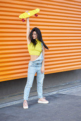 Beautiful active brunette girl with long hair wearing yellow top, blue denim overalls and stylish pink sneakers posing on orange wall background.