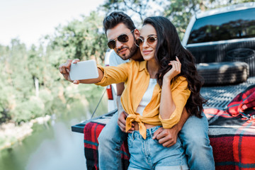 cheerful girl in sunglasses talking selfie with bearded man near car and lake