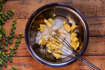 Metallic bowl with sugar and butter and a whisk preparing a cake on a wooden surface with green leaves