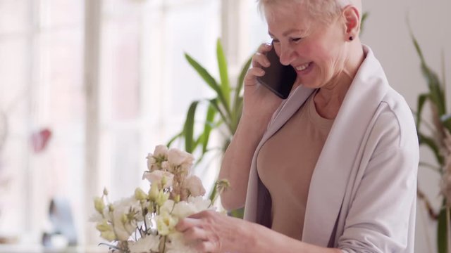 Tilt up of middle-aged Caucasian woman with short blonde hair standing at wooden table in workshop, smelling flowers and having talk with client using smartphone