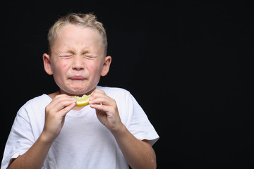 Little, blond boy is eating a piece of a lemon in front of black background and making a facial...
