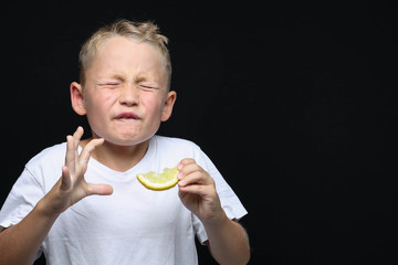 Little, blond boy is eating a piece of a lemon in front of black background and making a facial...